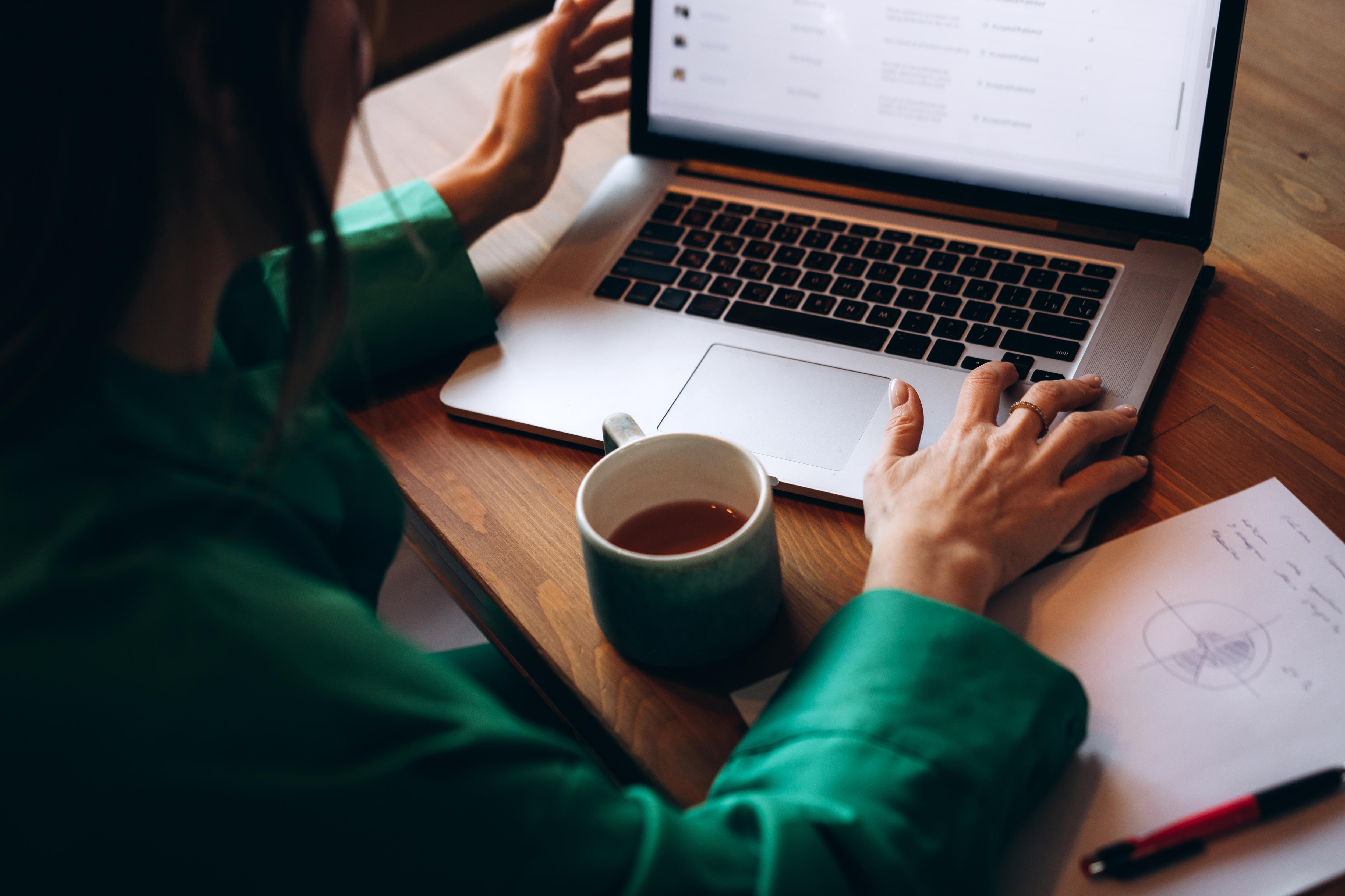 close up image of woman working on open laptop at table. Notes and coffee mug are also on table.