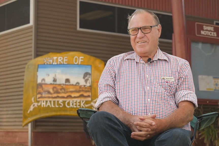A bespectacled man with thinning hair sits outside a building with a sign on it that reads "Shire of Halls Creek".