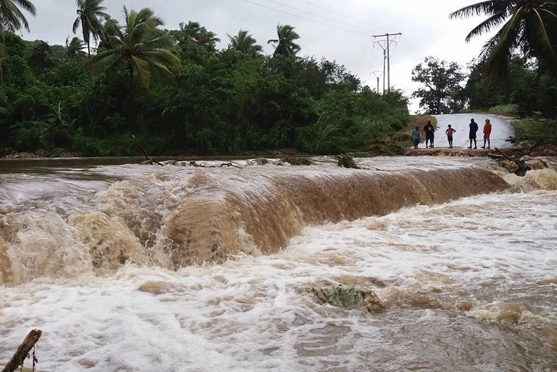 A group of people stand at the edge of a flooded road as rushes across.