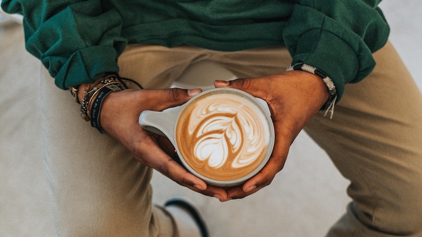 A photo of a man, shot from above, who's wearing a green shirt, beaded bracelets and holding a latte