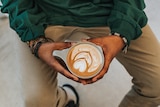 A photo of a man, shot from above, who's wearing a green shirt, beaded bracelets and holding a latte