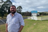 A man in a blue checked shirt stands smiling alongside a roadside and a fenced showground signposted 'Cobargo Showground'.