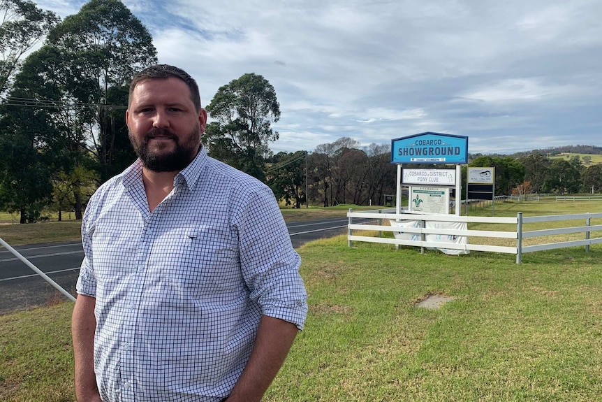 A man in a blue checked shirt stands smiling alongside a roadside and a fenced showground signposted 'Cobargo Showground'.