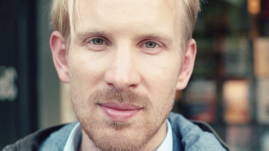 Close up photo of a white man with blonde hair wearing a green jacket and blue shirt
