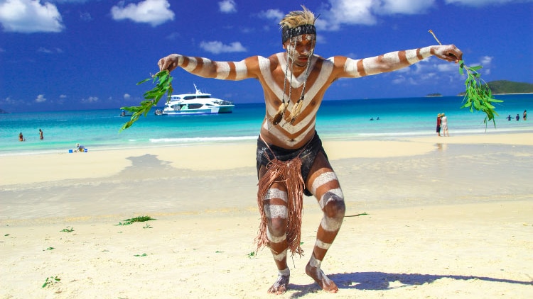 A young Indigenous man in traditional garb dances on Whitehaven Beach.