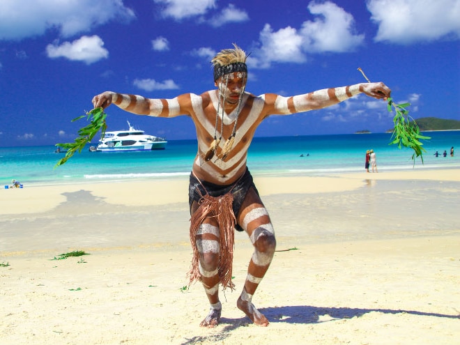 A young Indigenous man in traditional garb dances on Whitehaven Beach.