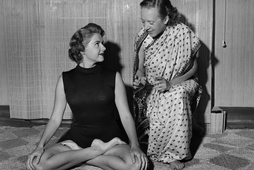 A black-and-white photo shows a woman in a sari instructing a yoga student.