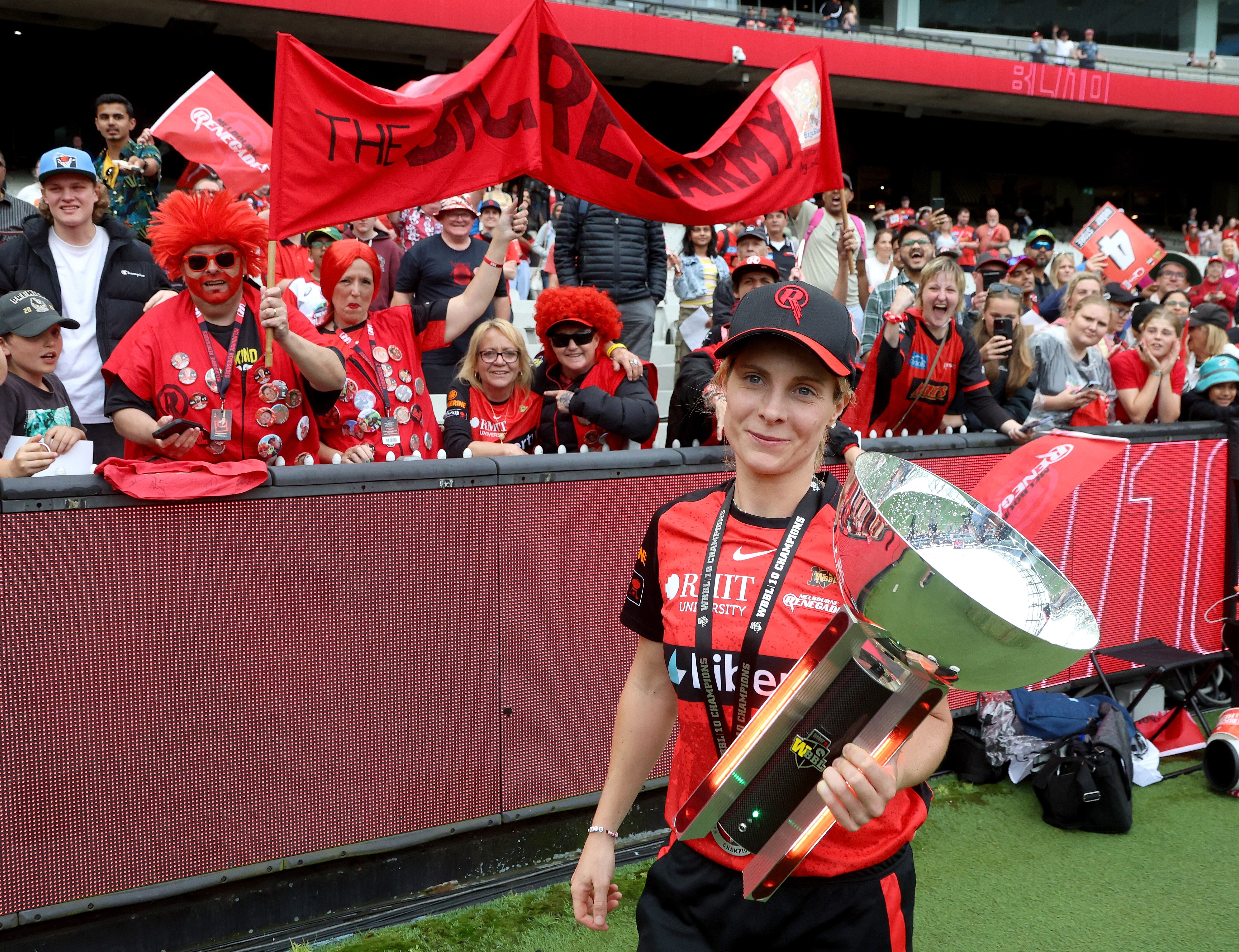 A Melbourne Renegades women's cricketer stands holding the WBBL trophy in front of a stand of fans after a final.