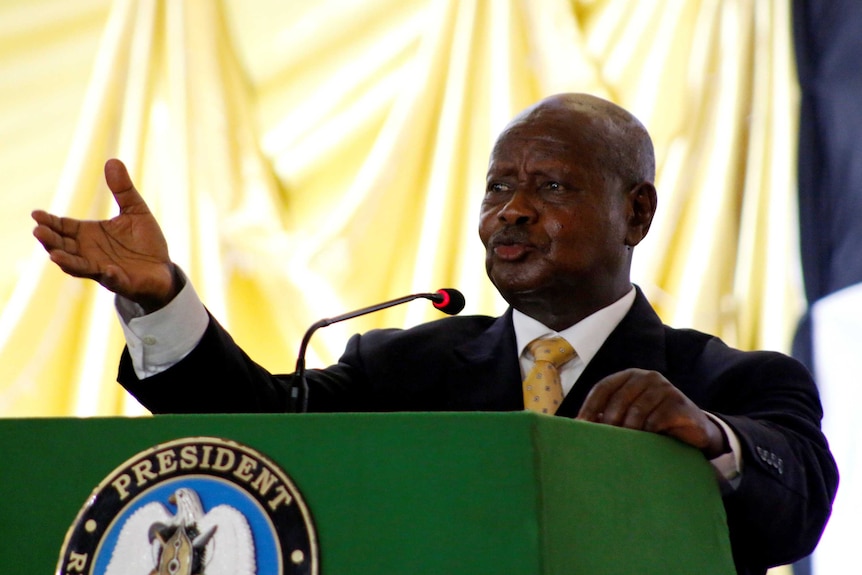 Ugandan President Yoweri Museveni holds his arm up as me speaks into a microphone behind a green lectern.