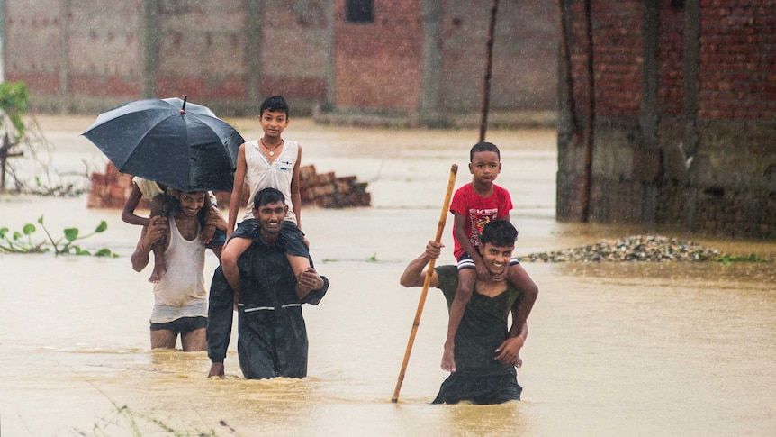 Nepalese men carry children on their shoulders in flood waters. The water is just above knee height.