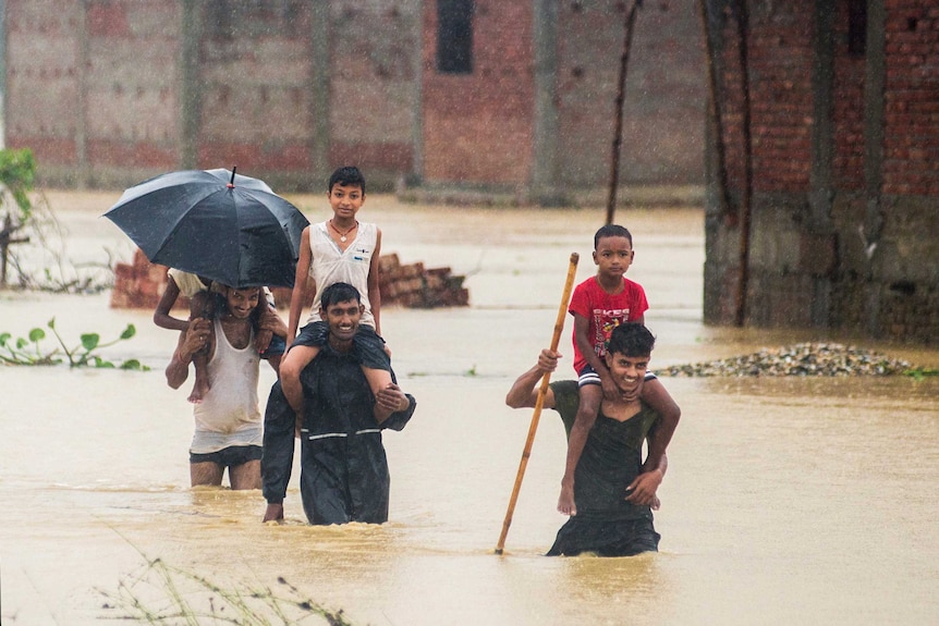 Nepalese men carry children on their shoulders in flood waters. The water is just above knee height.