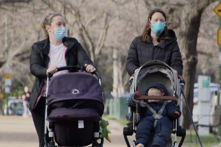 Two women wearing face masks push prams while walking in the gardens in Melbourne. One has a dog on a lead.