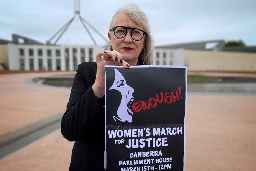 A woman is holding up a poster that says 'Enough' with details of a march, standing outside Parliament house in Canberra.