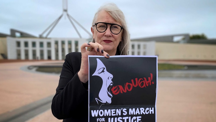 A woman is holding up a poster that says 'Enough' with details of a march, standing outside Parliament house in Canberra.