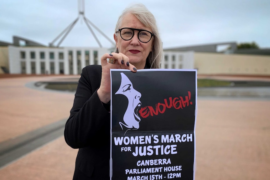 A woman is holding up a poster that says 'Enough' with the details of a march, standing outside the Parliament House in Canberra.