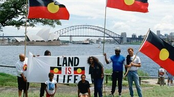 People hold Indigenous flags and "Land is Life" sign in front of Sydney Harbour, Opera House and Sydney Harbour Bridge