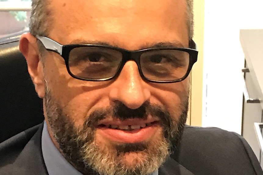 Close-up shot of a man with brown hair, greying facial hair, glasses and a smile.