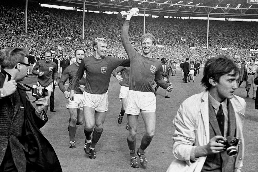 A black and white photo of Jack Charlton holding a trophy above his head at a packed Wembley