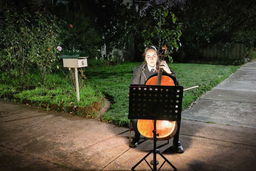 A girl in school uniform plays the cello while reading sheet music.