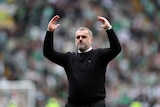 A football coach in his 50s with white hair and beard and black jumper raises his arms in celebration with blurred crowd behind 