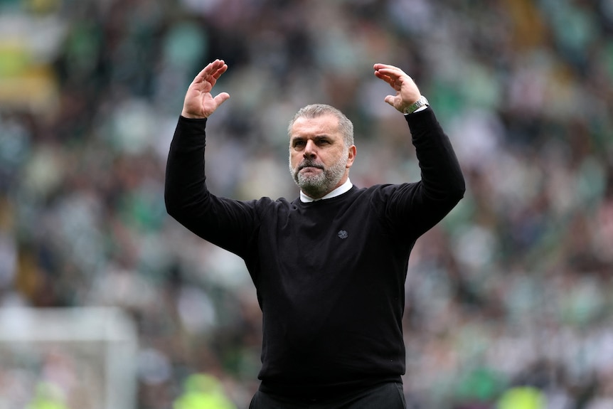 A football coach in his 50s with white hair and beard and black jumper raises his arms in celebration with blurred crowd behind 
