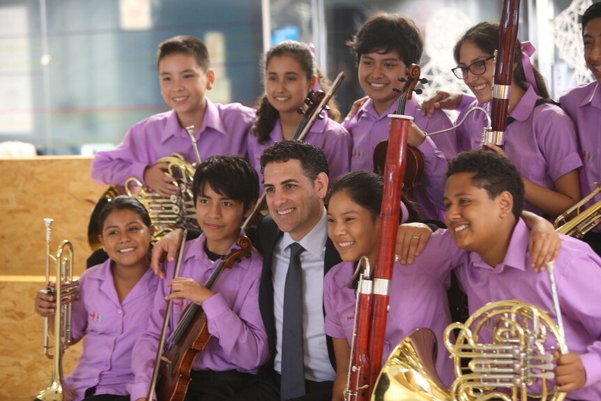 Juan Diego Flórez sits surrounded by young musicians holding instruments and wearing purple shirts.