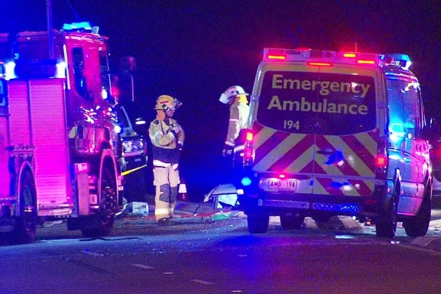 A fire truck and an ambulance on a road with a black sky and two emergency services crew in the middle