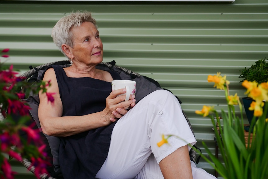 woman in mid-60s wearing navy top and white pants with white short hair sits in chair surrounded by flowers holding tea 