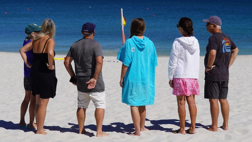 A shot from behind of a group of concerned locals standing in a line on Mullaloo Beach.