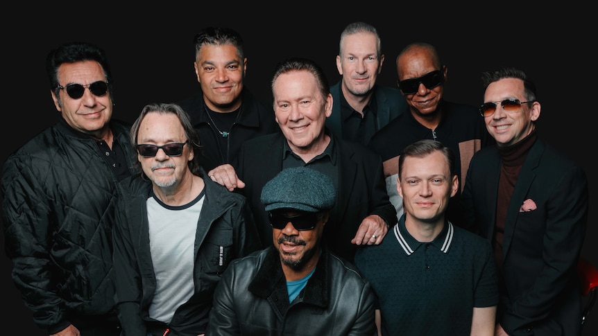 Black background with nine men from waist up huddled together smiling at camera. 