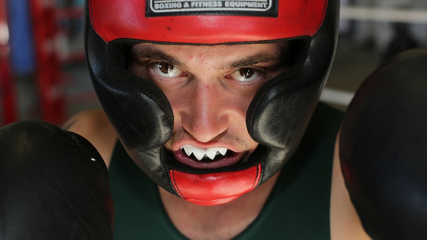 A boxer wears a red and black helmet as well as a white mouthguard and looks directly into the camera with gloves.