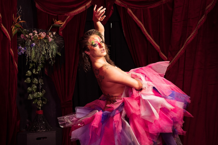 White woman with wet brown hair and pink, gold and green face paint strikes a dance pose, wearing pink tulle on a stage.