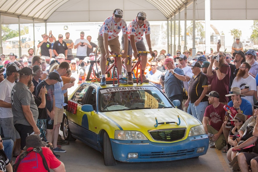 Two men sit on bikes stuck to the roof of a yellow and blue car. There is a large crowd around them.