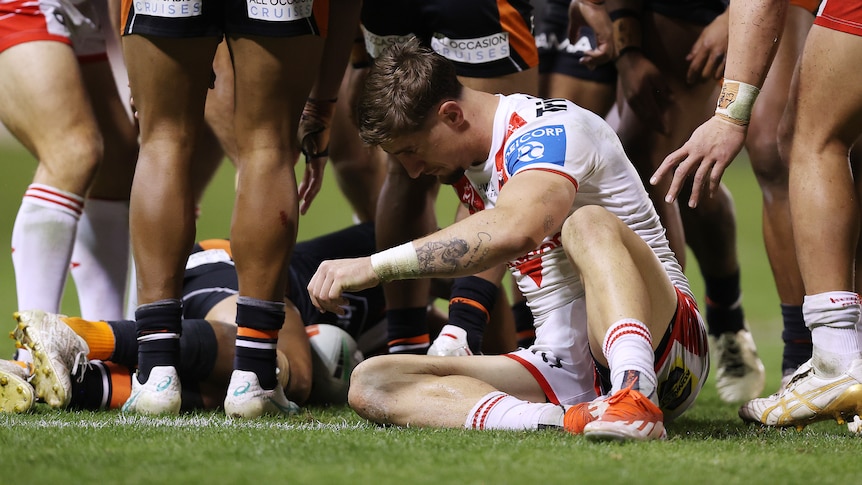 An NRL player sitting on the ground groggy, while opponents surround a fallen player in the backfield.
