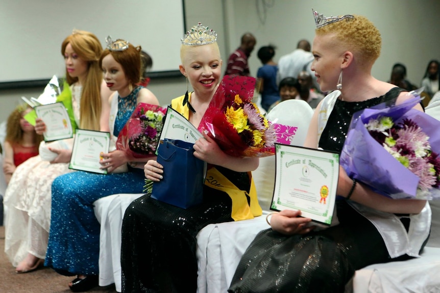 In a dimly-lit conference room, a group of four albino pageant contestants sit holding certificates and flower bouquets.