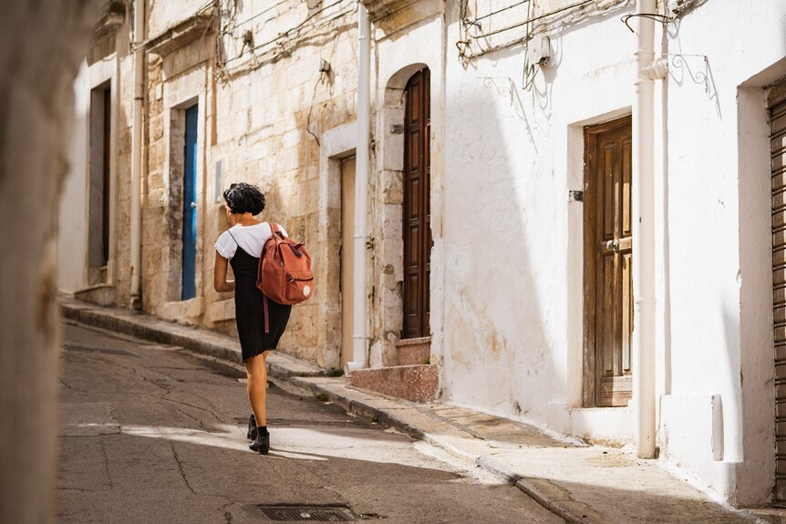 Carrying a small backpack, Lisa walks uphill through the narrow street of an old European city 