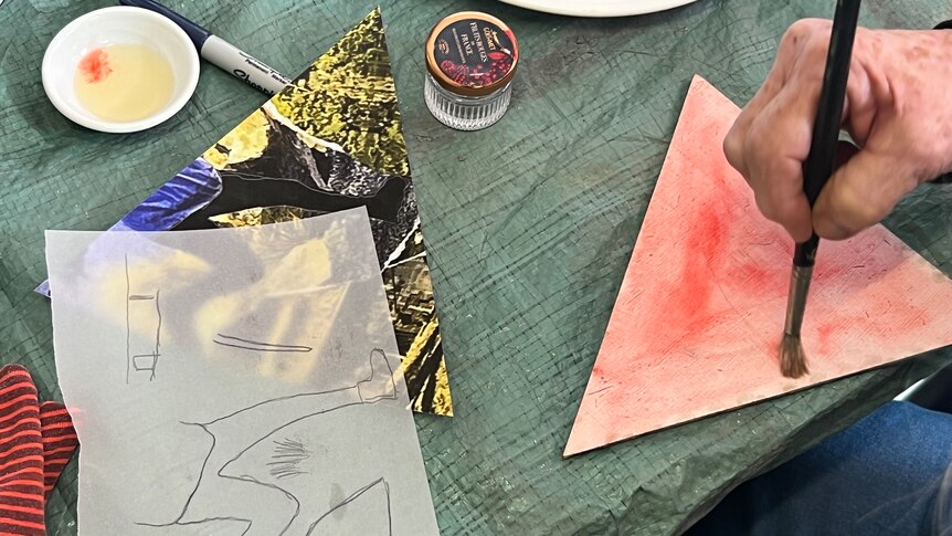 Tracing paper, watercolour paint and brushes are scattered across an art table, a hand works some opaque red paint into paper