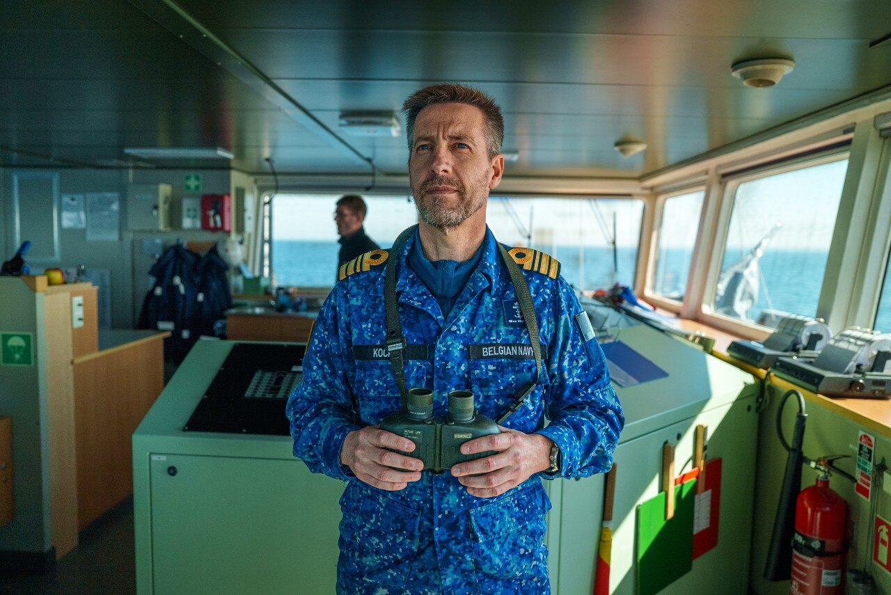 A man pictured inside the bridge of a ship, with his hands holding binoculars, looking on.