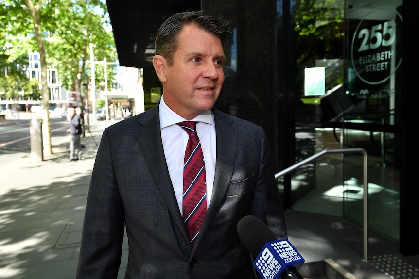 A man in a suit and tie walks past as a microphone is held out towards him. 