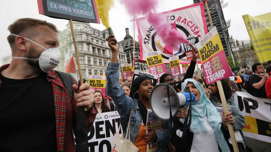 Woman in headscarf speaks in megaphone as people carry signs and send yellow and pink flares in the air