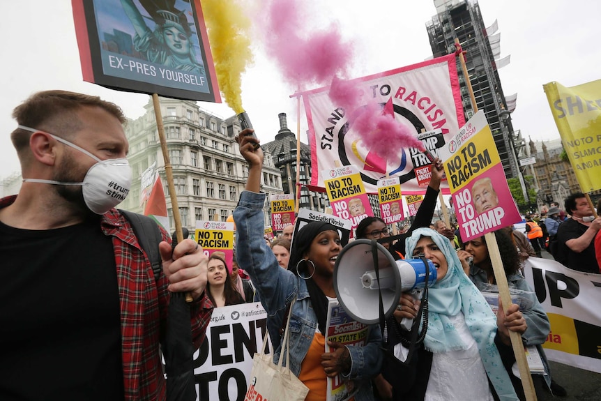 Woman in headscarf speaks in megaphone as people carry signs and send yellow and pink flares in the air