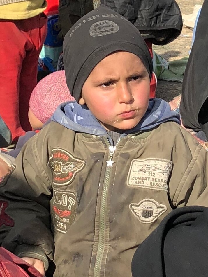 A young boy wears an IS beanie while sitting with other children in an open area