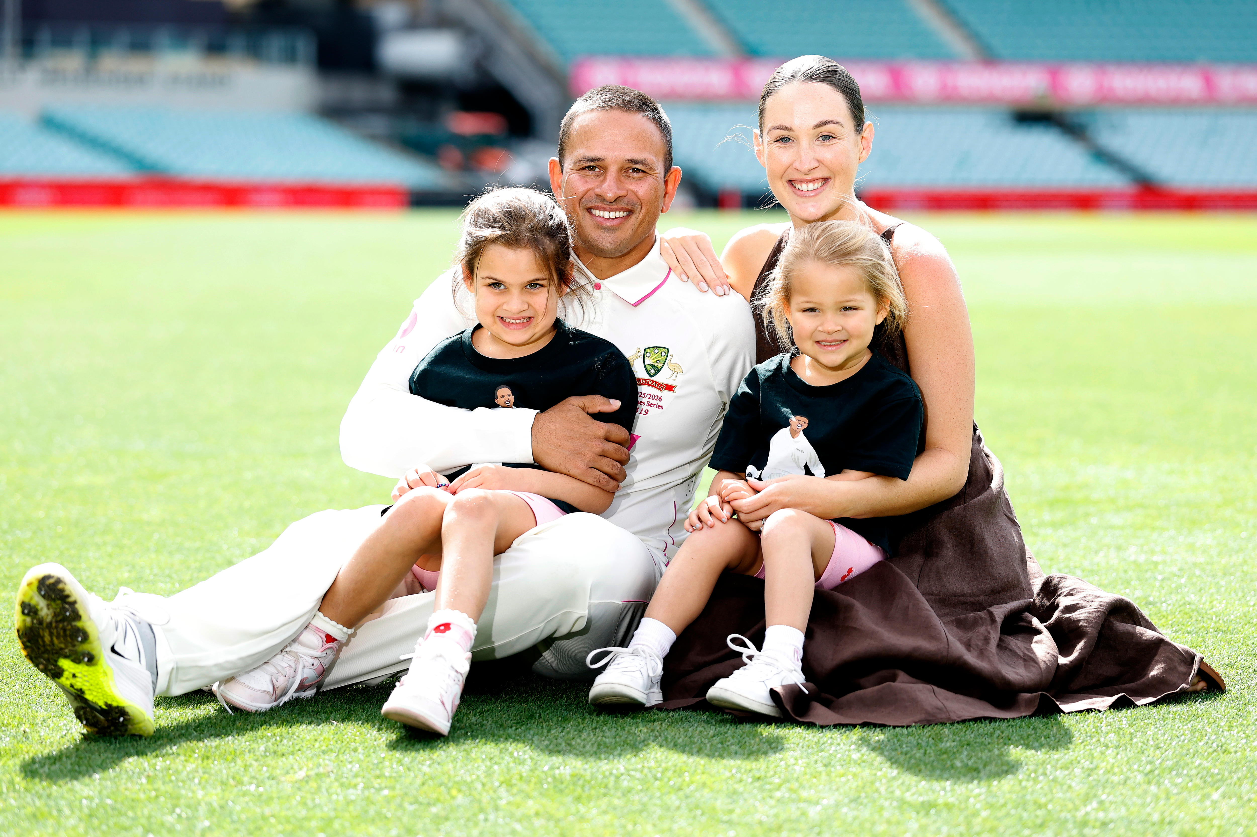 Usman and Rachel Khawaja smile big while seated on the cricket field grass with two young daughters on their laps.
