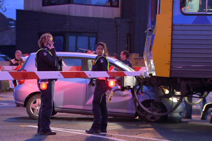 Two female police officers stand in front of the wreck of a car which is under a train.