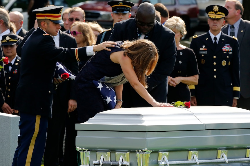 A woman bent over a coffin while a man in dress military uniform comforts her
