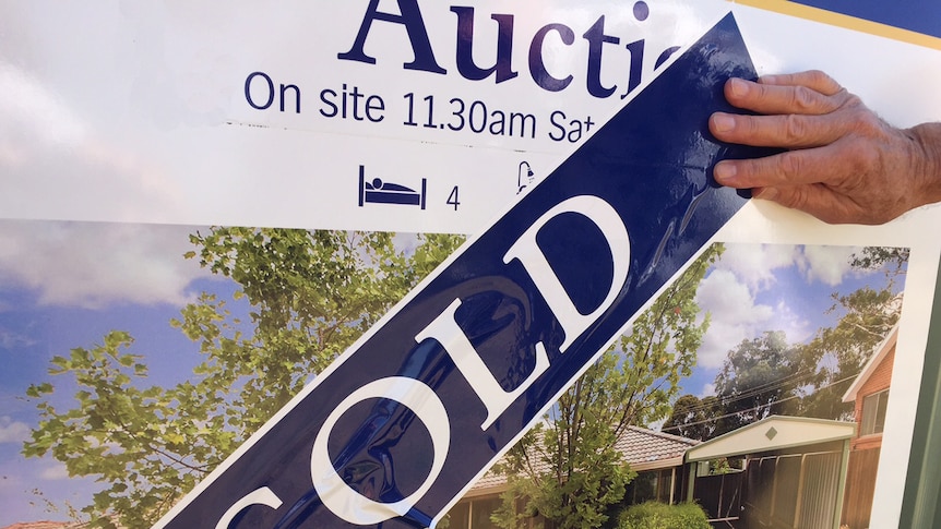 A hand smooths out a 'SOLD' banner over an auction billboard for a home.