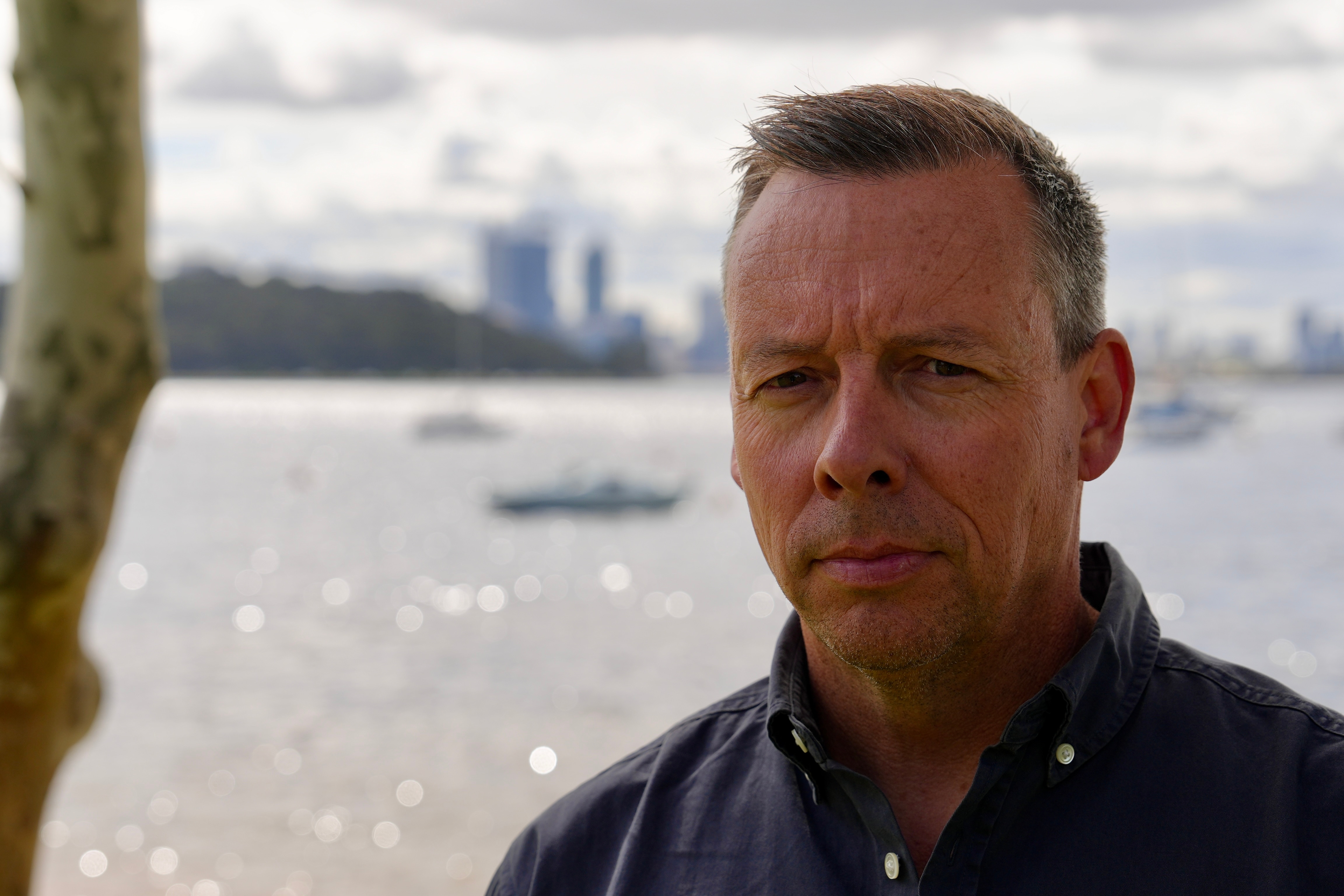 A man looks straight at the camera with a serious expression with the sparkling waters of the Swan River behind him.