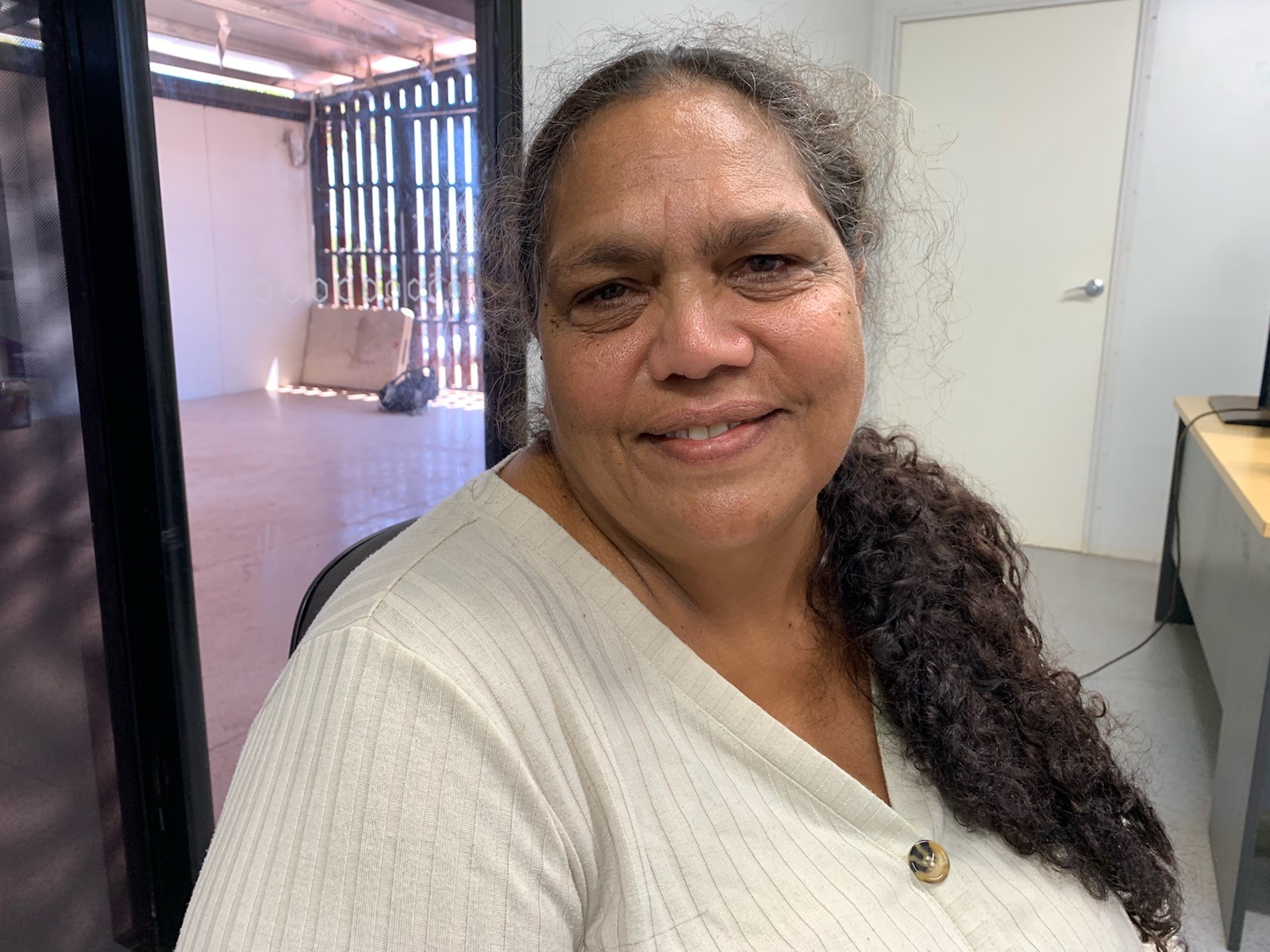 Aboriginal woman sitting on office chair, long curly hair on side pony-tail, sitting in office, smiling at camera.