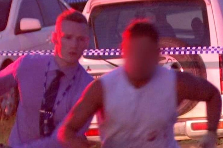 A man escapes a police officer and runs towards the camera with police cars in the background.