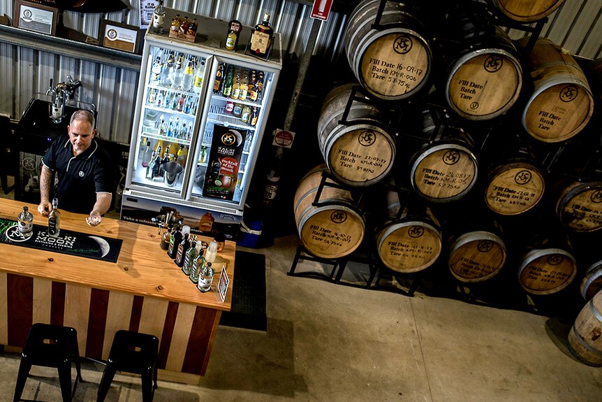 Barrels and a bar in a distillery shop front and man serves drink.